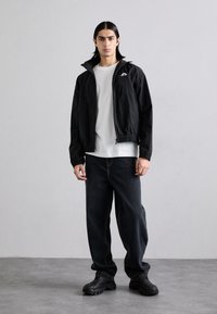 Black lightweight jacket with elastic waistband, worn over a white shirt. Paired with dark blue jeans and black textured sneakers.
