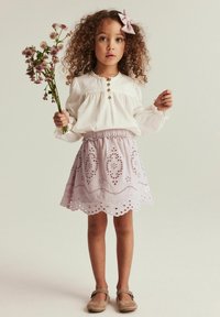 Helles rosa Kleid mit gewelltem Saum und bestickten Mustern, kombiniert mit einer weißen Bluse mit langen Ärmeln, die drei Knöpfe und Spitzenakzente aufweist.