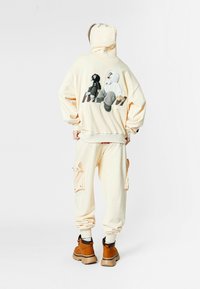 Persona con sudadera beige y pantalones cargo con figuras en blanco y negro al estilo de dibujos animados en la parte trasera de la sudadera, llevando botas marrones, sobre un fondo blanco.