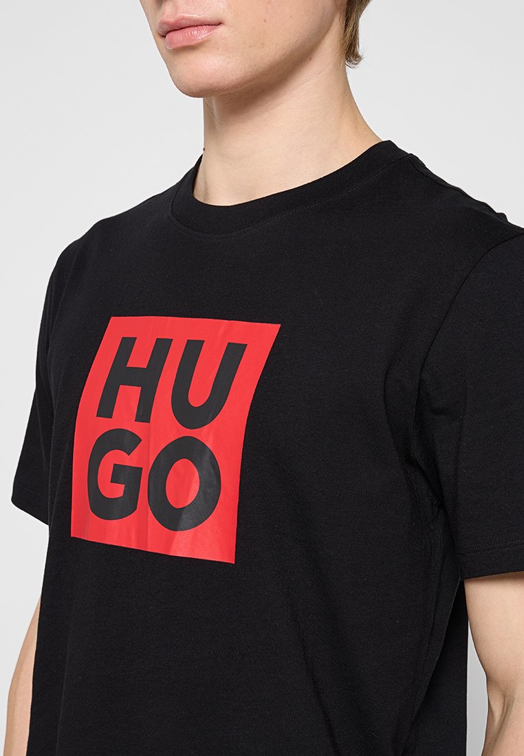 Camiseta de algodón negra con un gran cuadrado rojo que presenta en el centro un texto en negrita de color negro que dice "HUGO". Cuello redondo y mangas cortas.