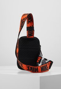 Černá síťovaná taška přes rameno s nastavitelním oranžovým popruhem a černým logem Nike, obsahující zapínací kapsu.