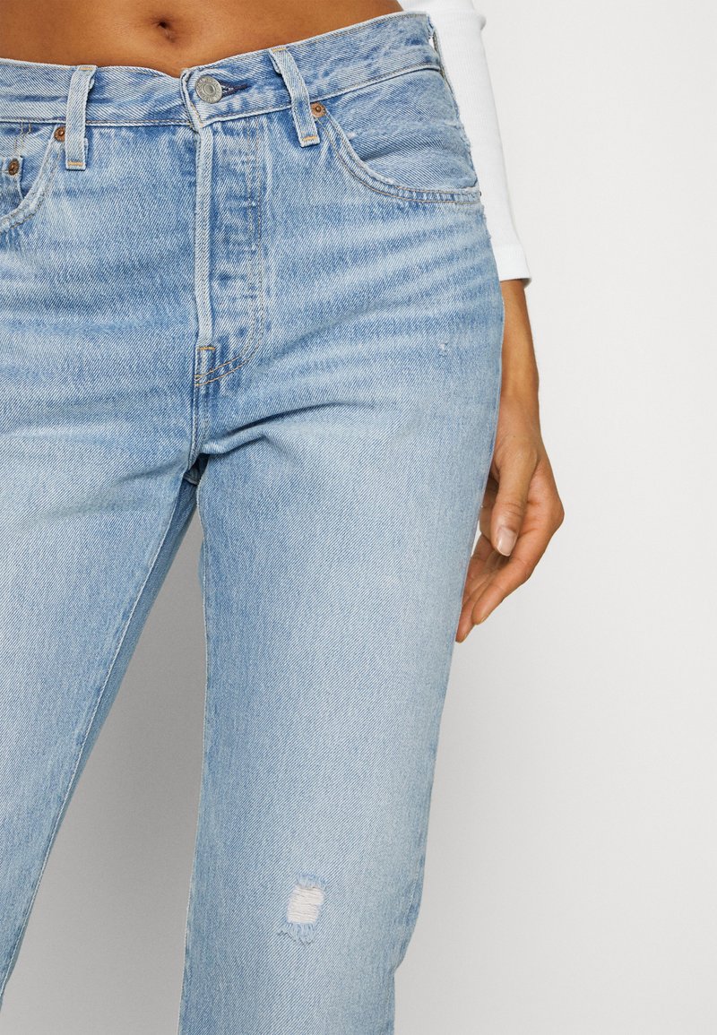 Vaqueros de denim azul claro con cintura alta, diseño de cinco bolsillos y detalles desgastados en el muslo izquierdo. Desgaste sutil en todo el tejido.