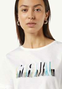 Weißes Baumwoll-T-Shirt mit mehrfarbigem, gesticktem Text "CASUAL" und Fransenverzierungen. Runder Ausschnitt und kurze Ärmel.