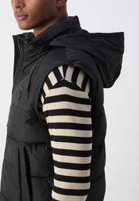 Black puffer vest with a hood, featuring quilted fabric and a zipper closure. Underneath, a striped long-sleeve shirt in beige and black.