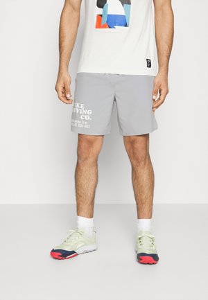 Hombre de pie con pantalones cortos grises con texto blanco, calcetines blancos, zapatillas verde claro y rojas, y una camiseta blanca con un diseño geométrico colorido.