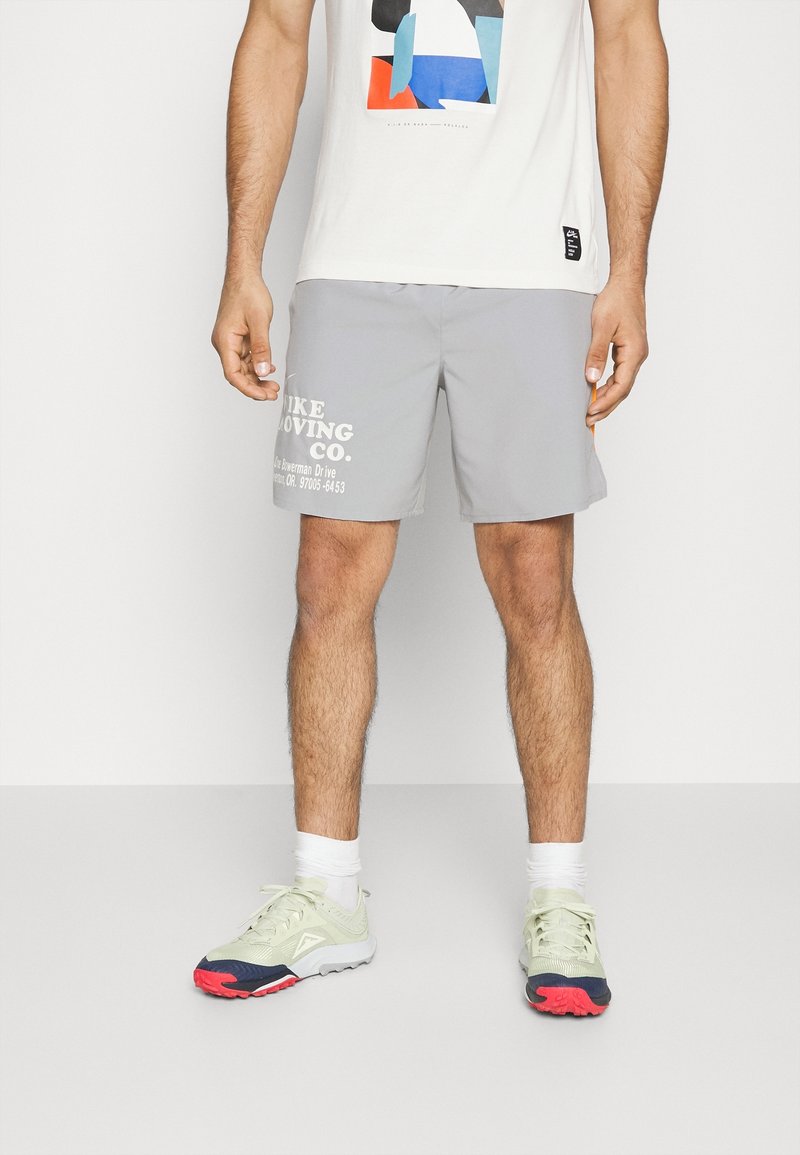 Homme debout portant un short gris avec un texte blanc, des chaussettes blanches, des baskets vert clair et rouges, et un t-shirt blanc avec un motif géométrique coloré.