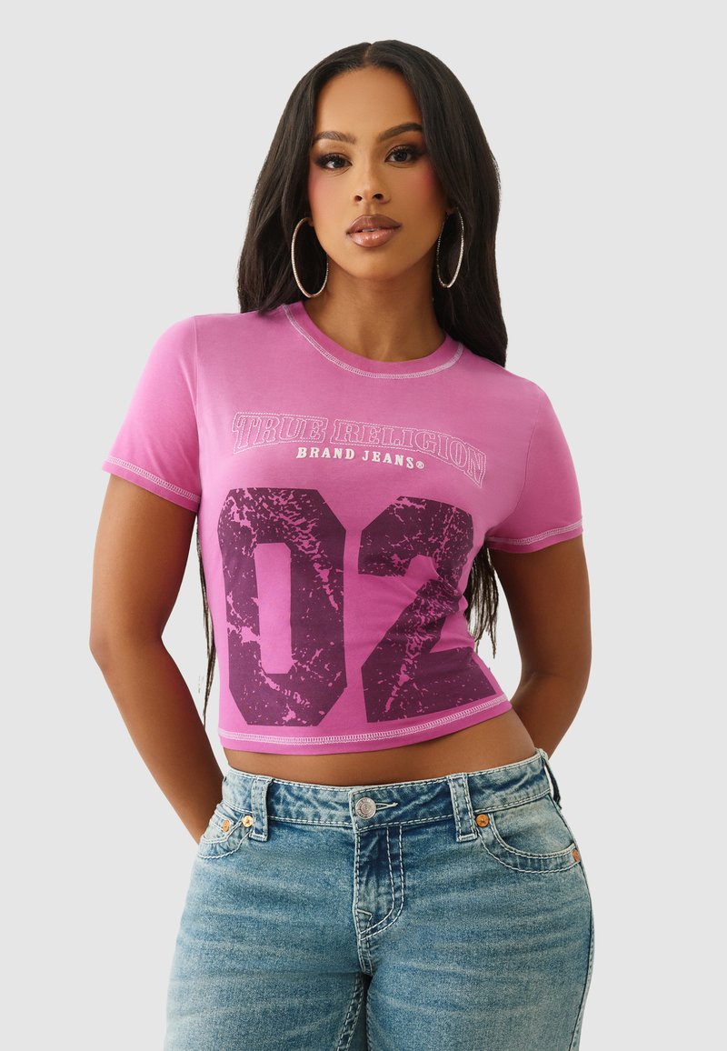 Różowy krótki top z krótkim rękawem z napisem „TRUE RELIGION” i dużą grafiką „02” w ciemnofioletowym kolorze. Gładka tekstura i kontrastowe detale przeszyć.