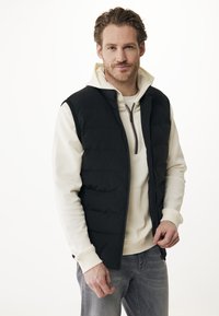 Muž s světlohnědými vlasy a plnovousem, oblečený v krémové mikině a černé péřové vestě, stojící na jednobarevném bílém pozadí.