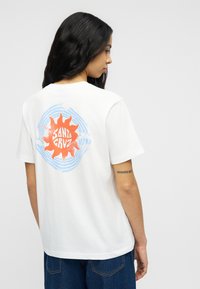 Γυναίκα με μακριά μαύρα μαλλιά, που φοράει λευκό μπλουζάκι με πορτοκαλί ήλιο και το κείμενο "Santa Cruz" σε μπλε κυκλικό φόντο στην πλάτη.