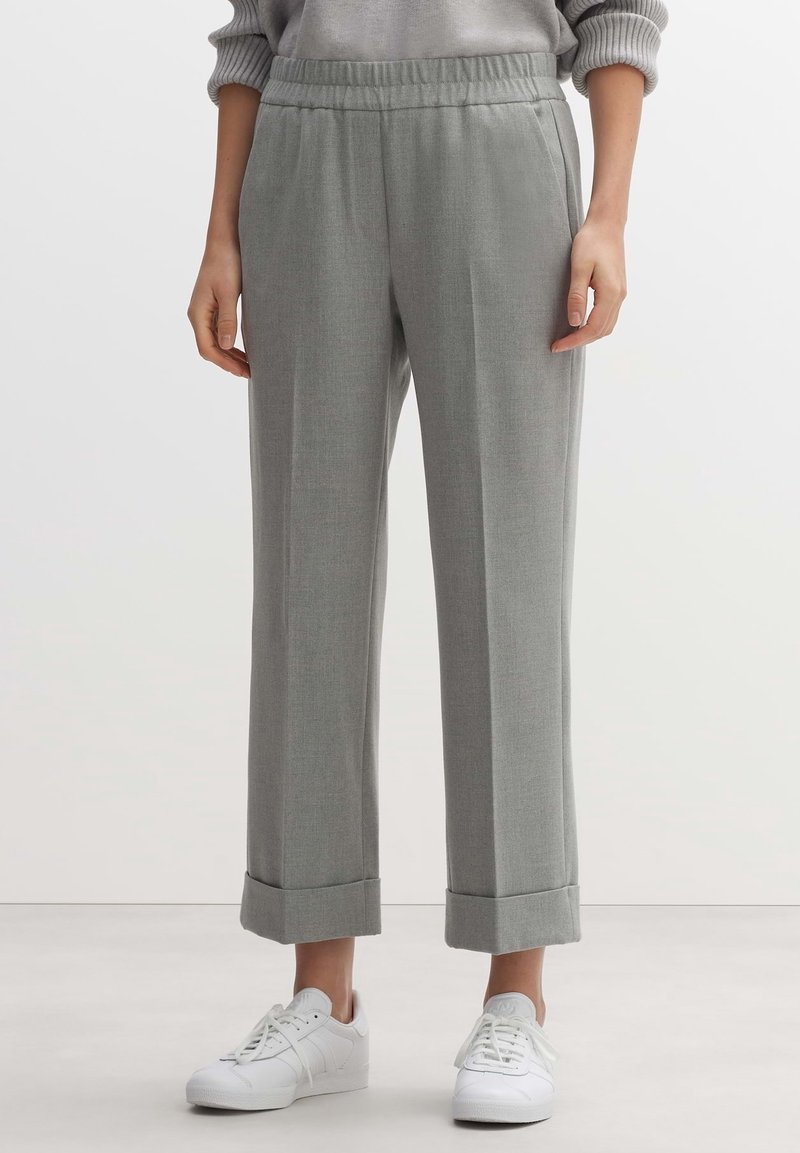 Personne portant un pantalon large gris clair avec des ourlets retroussés et une taille élastique, associé à des baskets blanches, debout sur un sol clair.