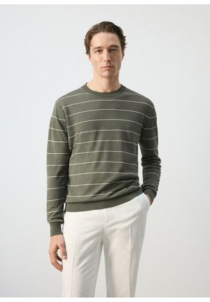 Hombre con suéter de manga larga color verde oliva con finas rayas horizontales blancas y pantalones blancos, de pie con una mano en el bolsillo.