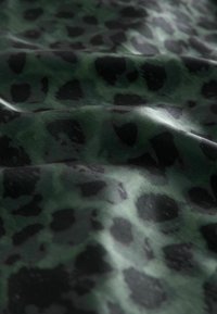 Zaļa un melna rakstaina auduma ar gludu tekstūru, kas satur neregulāras leopardu plankumus dažādās formās un izmēros.