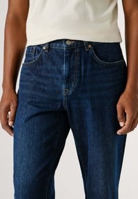 Jeans de denim azul oscuro con un diseño de pierna recta, adornados con costuras naranjas, estilo de cinco bolsillos y un cierre de botón metálico en la cintura.