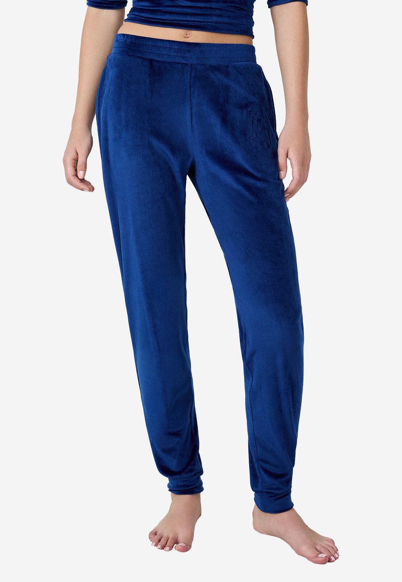 Kobieta w niebieskich dresowych pantalonach z elastycznym pasem i ściągaczami przy kostkach, stojąca boso na białym tle.