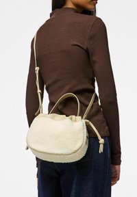Borsa beige e crema realizzata in materiale shearling testurizzato. Forma morbida e arrotondata con parte superiore a cordoncino e tracolla regolabile.