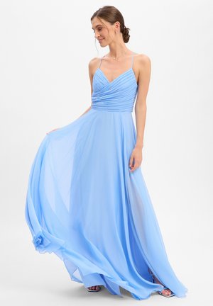 Frau in einem fließenden hellblauen ärmellosen Kleid mit dünnen Trägern, die den Rock hält und mit tief gestecktem Haar nach unten schaut.