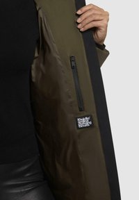 Olivgrüne Jacke mit schwarzem Futter, ausgestattet mit einer Reißverschlusstasche und einem Markenetikett "Khujo" auf der Innenseite. Glatte Textur, zeitgemäßes Design.