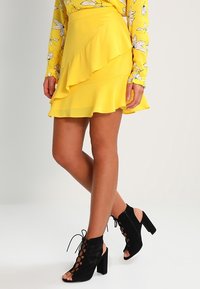 Gelbe, mehrlagige Minirock mit volantren Saum, kombiniert mit schwarzen Wildleder-Open-Toe-Absätzen mit Knöchelriemchen und Blockabsätzen.