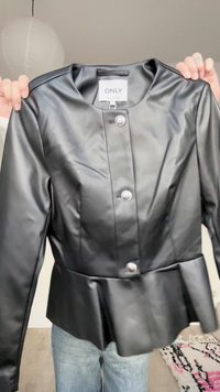 Μαύρο μπουφάν από ψεύτικο δέρμα με πεπλουμ τελείωμα και τρία ασημένια κουμπιά, κρατημένο από χέρια μπροστά σε φωτεινό φόντο.