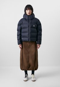 Navy puffer jacket with a high neck, featuring quilted sections and a logo badge. Worn over a brown, knee-length skirt and white sneakers.