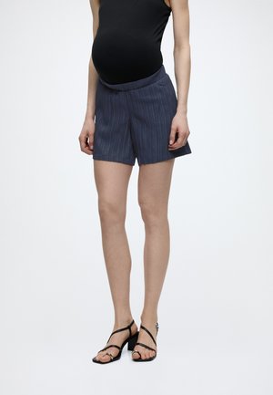 Έγκυος που φοράει μαύρο αμάνικο τοπ, σκούρο μπλε ριγέ σορτς εγκυμοσύνης και μαύρα πέδιλα με τακούνι και λουριά, στέκεται μπροστά σε λευκό φόντο.