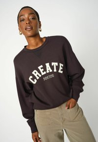 Tmavofialová mikina s veľkým bielym nápisom "CREATE" a "MM2010" pod ním. Dlhé rukávy a voľný strih v kombinácii s béžovými nohavicami.