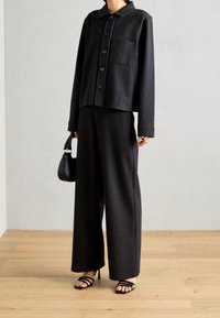 Giacca su misura nera con righe sottili, caratterizzata da grandi bottoni e una tasca sul petto, abbinata a pantaloni larghi coordinati. Presenta una texture liscia.