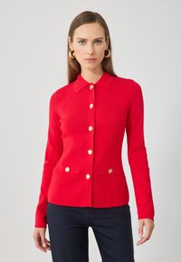 Cárdigan de punto rojo con cuello, con botones dorados y dos bolsillos frontales. Corte ajustado y mangas largas. Textura suave.