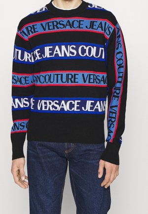 Mann trägt schwarzen Pullover mit horizontalen blauen, roten und weißen Streifen und dem Schriftzug "Versace Jeans Couture", kombiniert mit dunkelblauen Jeans.
