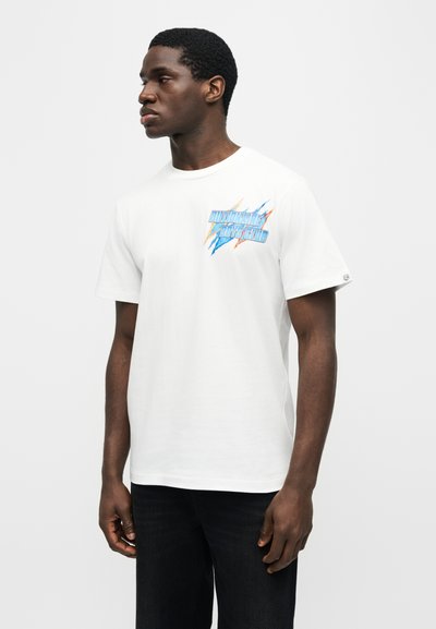 Νεαρός άνδρας που φοράει λευκό μπλουζάκι με πολύχρωμο λογότυπο «Billionaire Boys Club» στο στήθος, στέκεται μπροστά σε απλό φωτεινό φόντο.