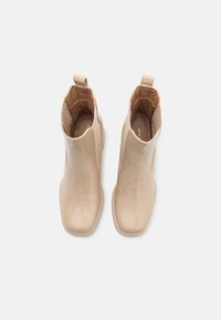 Beżowe botki do kostki z błyszczącego lakierowanego wykończenia, kwadratowym noskiem i elastycznymi panelami bocznymi. Posiadają pętelkę z tyłu ułatwiającą zakładanie.