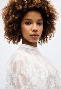 Νεαρή γυναίκα με καστανά σγουρά μαλλιά, φορώντας ασημένια σκουλαρίκια κρίκους και λευκό διάφανο κορυφαίο με δαντέλα και λουλούδια, κοιτάζοντας με αυτοπεπο ίθηση την κάμερα.