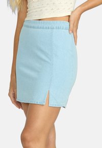 Falda corta de mezclilla azul claro con una abertura frontal, dobladillo sin terminar y cinturilla elástica. Textura suave con una silueta ajustada.