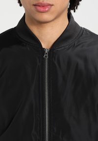 Schwarze Bomberjacke mit Satin-Finish, ausgestattet mit einem Frontreißverschluss, einem gerippten Kragen und sichtbaren Steppnähten.