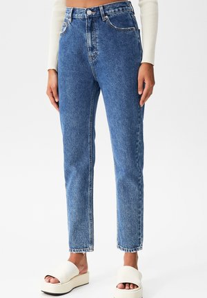 Frau trägt hoch taillierte blaue Jeans mit geradem Bein und weiße Plateau-Sandalen, steht vor einem schlichten weißen Hintergrund.