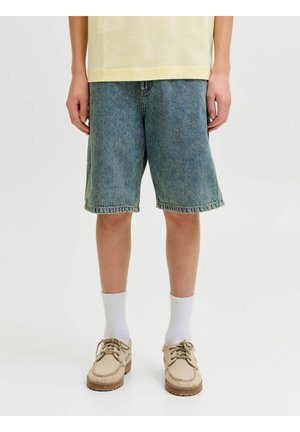 Person iført løse blå denimshorts, hvite crew-sokker og beige snøresko, stående mot en ensfarget lys bakgrunn.