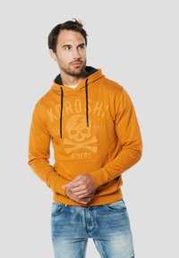 Narancssárga kapucnis pulóver fekete húzózsinórokkal, koponya és csontváz motívummal. Puha tapintású, kenguruzsebbel és bordázott mandzsettákkal.