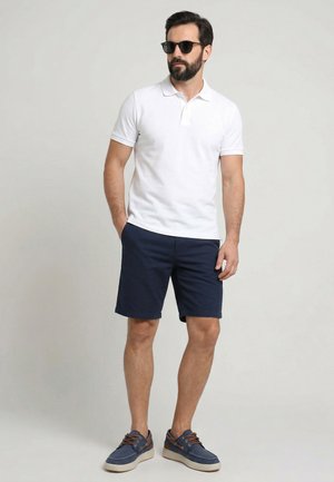 Mann trägt schwarze Sonnenbrille, weißes kurzärmeliges Poloshirt, marineblaue Shorts und blaue Bootsschuhe, steht mit einer Hand in der Tasche.