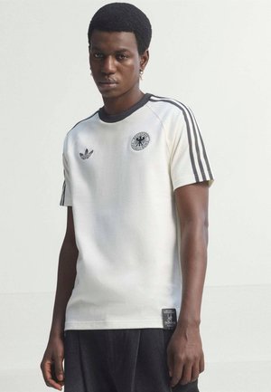 Mladý muž oblečený v bielom futbalovom tričku Adidas s čiernymi pruhmi a logom nemeckého národného tímu stojaci pred jednofarebným pozadím.
