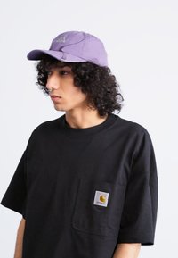 Purple cotton cap with curved brim and embroidered logo, paired with a loose-fitting black t-shirt featuring a front pocket and logo patch.