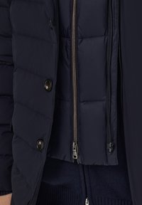 Jūras spēku pūkainā jaka ar quilted tekstūru, priekšējo rāvējslēdzēju un uzspiežamajiem pogām, ar slāņainu iekšējo dizainu un kontrastējošu audumu.