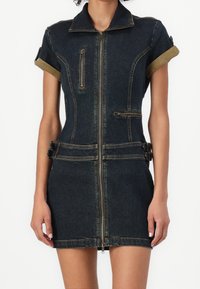 Trafikpassad mörk denim mini-klänning med framzip, uppvikta korta ärmar, dekorativa zipfickor och sidospännen på en stående person.
