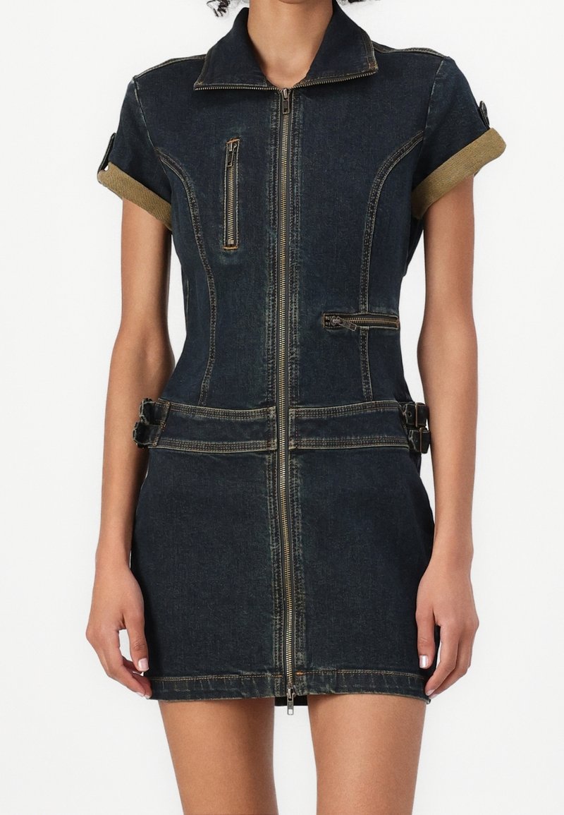 Trafikpassad mörk denim mini-klänning med framzip, uppvikta korta ärmar, dekorativa zipfickor och sidospännen på en stående person.