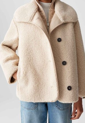 Beige sherpa fleecejack met grote kraag, zwarte knopen en zijzakken, gedragen over een beige trui met rits en wit overhemd, gecombineerd met blauwe jeans.