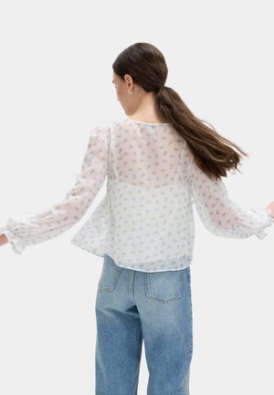 Mujer con cabello largo recogido en una coleta baja, vistiendo una blusa blanca translúcida con estampado floral azul y unos vaqueros de talle alto en azul claro, de espaldas.