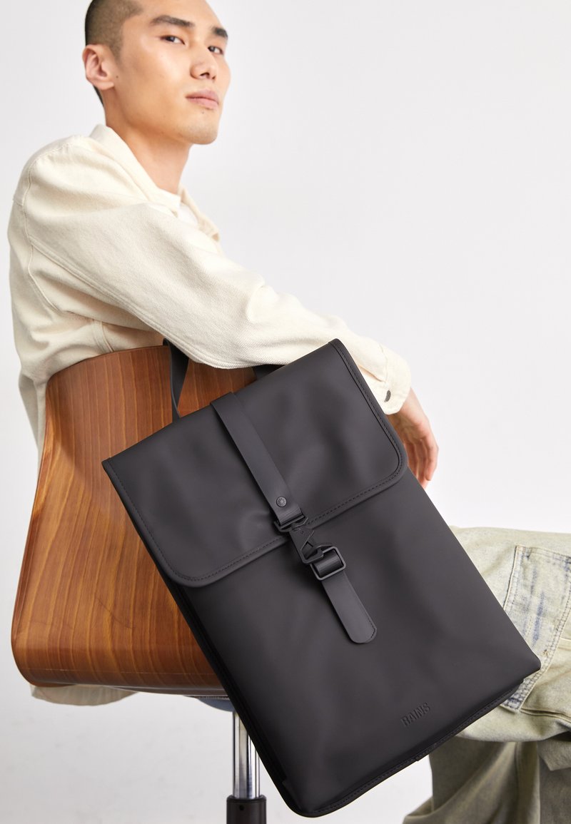 Schwarzer wasserdichter Rucksack mit Klappenverschluss, Schnallen-Details und minimalistischem Design. Glatte Textur mit dem eingravierten Markennamen "RAINS".