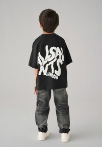 Černé oversized tričko s bílým stylizovaným nápisem "ALLSAINTS" na zadní straně, spárované se šedými džíny a bílými teniskami.