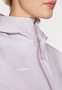 Šviesiai levandų spalvos nepralaidi vandeniui striukė su aukštu apykakle, turinti priekinį užtrauktuką ir reguliuojamą kapisoną. Ant krūtinės yra "Mammut" logotipas.