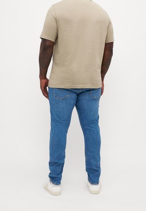 Beige Baumwoll-T-Shirt und blaue Denim-Jeans. Das Shirt hat einen Rundhalsausschnitt; die Jeans sind eng geschnitten, mit Gesäßtaschen und geradem Schnitt.