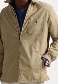 Béžová zipová bunda s límcem, přední kapsou a vyšitým logem. Nositá přes modrou košili, včetně viditelného prošívání a volného střihu.